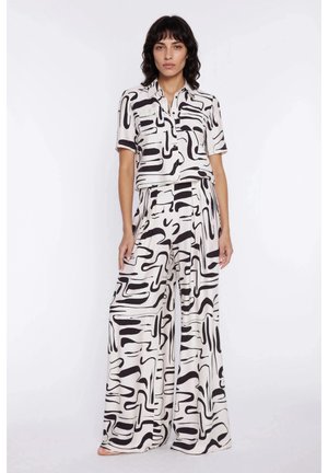 Kobieta stojąca w pasującej czarno-białej koszuli z krótkim rękawem z abstrakcyjnym wzorem i szerokich spodniach na jednolitym tle.