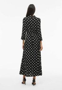 Černé šaty s puntíky, tříčtvrteční rukávy, vypasovaný pas a rozšířená sukně. Hedvábná látka, délka po kotníky, v kombinaci s černými podpatky.