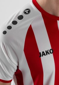 Maglietta a righe rosse e bianche realizzata in tessuto sportivo. Presenta accenti circolari neri sulle spalle e il logo "JAKO" sul davanti.