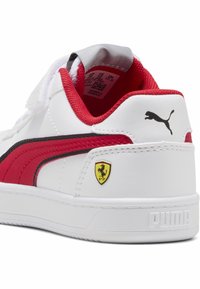 Weißer Sneaker mit roten Akzenten, ausgestattet mit einem schwarzen Streifen und dem Ferrari-Logo. Hergestellt aus glattem synthetischem Material mit einer strukturierten Zunge und gepolstertem Kragen.
