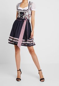 Femme portant une robe traditionnelle dirndl avec un chemisier en dentelle, un bustier à motifs, une jupe bleu marine, un tablier rose noué à la taille et des sandales à talons hauts noires.
