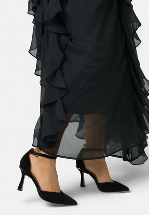 Mujer con zapatos de tacón negro de punta afilada y tiras en el tobillo, y una falda midi negra transparente con volantes.