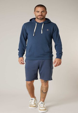 Mann mit kurzen Haaren und Bart, trägt einen blauen Hoodie, passende Shorts und creme-weiße Turnschuhe, steht vor einem einfarbigen Hintergrund.