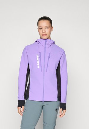 Kvinne som har på seg en lavendel fôret hettegenser med svarte sidelapper og grå bukser, stående mot en enkel hvit bakgrunn.