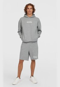 Grijze hoodie en shortset, gemaakt van zacht materiaal, met een vo zak en witte logotekst, gecombineerd met witte sneakers.