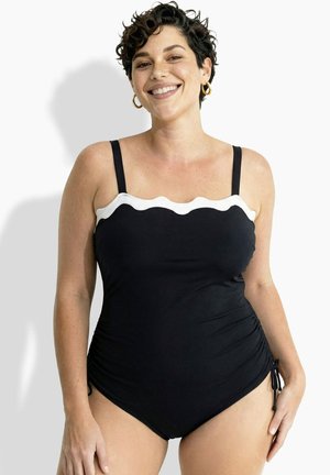 Donna sorridente con capelli corti e ricci, che indossa un costume intero nero con bordatura ondulata bianca e orecchini a cerchio dorati, su sfondo bianco.