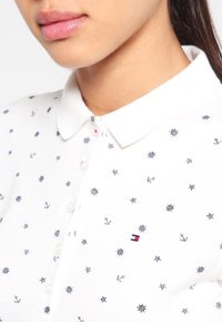 Bílá polo tričko s rozptýlenými námořními vzory v námořnické modři, s límečkem, knoflíkovou légou a malým vyšitým logem poblíž hrudi.