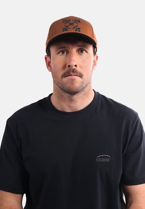 Casquette de baseball marron avec un design brodé foncé, portée avec un t-shirt noir. La casquette présente une visière structurée et un dos ajustable.