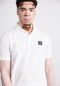 Balts polo krekls, izgatavots no kokvilnas, ar klasisku apkakli un melnu Timberland logo uzšuvumu kreisajā pusē. Īsas piedurknes un gluda tekstūra.