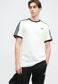 Biele tričko s čiernymi akcentmi a tromi čiernymi pruhmi na pleciach. Obsahuje malé čierne logo na ľavom hrudi. Hladká bavlnená textúra.