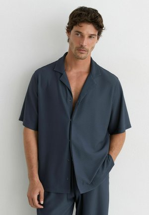 Man wearing textured dark blue short-sleeve shirt and matching shorts, standing with one hand in pocket against a white wall.