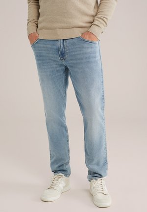Persona vistiendo jeans de color azul claro, suéter beige y zapatillas blancas, de pie con las manos en los bolsillos frente a un fondo liso.