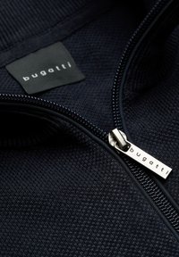 Marineblaue Jacke mit Struktur und teilweise geöffnetem Reißverschluss, der einen Metall-Zipper und ein inneres Etikett zeigt, beide mit der Marke "bugatti".