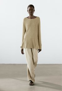 Maglione a coste beige con scollatura ampia, abbinato a pantaloni a gamba larga beige chiaro e sandali neri, il tutto su uno sfondo minimalista.