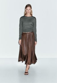 Vrouw loopt zelfverzekerd in een grijze lange mouwen top, een bruine geplooide midirok, zwarte sandalen met bandjes en zilveren creolen.