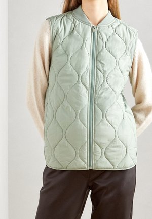 Gilet sans manches matelassé vert clair avec un motif de vagues texturé, fermeture éclair sur le devant et col côtelé. Porté par-dessus un haut à manches longues beige.