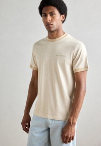 Beige bavlněné tričko s krátkými rukávy, kontrastními manžetami a límcem. Na hrudi je jemně tištěné logo značky v světle hnědé barvě.