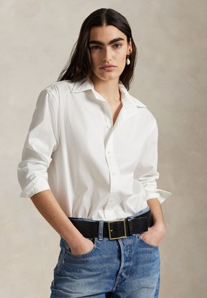 Pitkähiuksinen nainen, jolla on valkoinen napillinen paita, siniset farkut mustalla vyöllä ja korvakorut, poseeraa käsillään taskuissa.