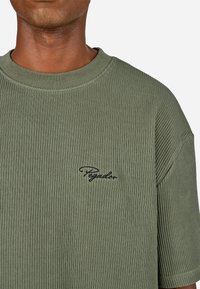 Alyvuogių žalios spalvos dryžuotas marškinėlis, pagamintas iš tekstūruotos medžiagos, su apvaliu iškirpimu ir mažu juodu siuvinėtu logotipu ant krūtinės.