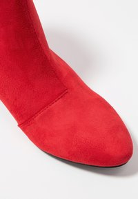 Czerwone zamszowe botki za kostkę z czubkiem w kształcie szpica i gładką teksturą, z bezszwowym designem i elegancką sylwetką. Widoczna czarna podeszwa.