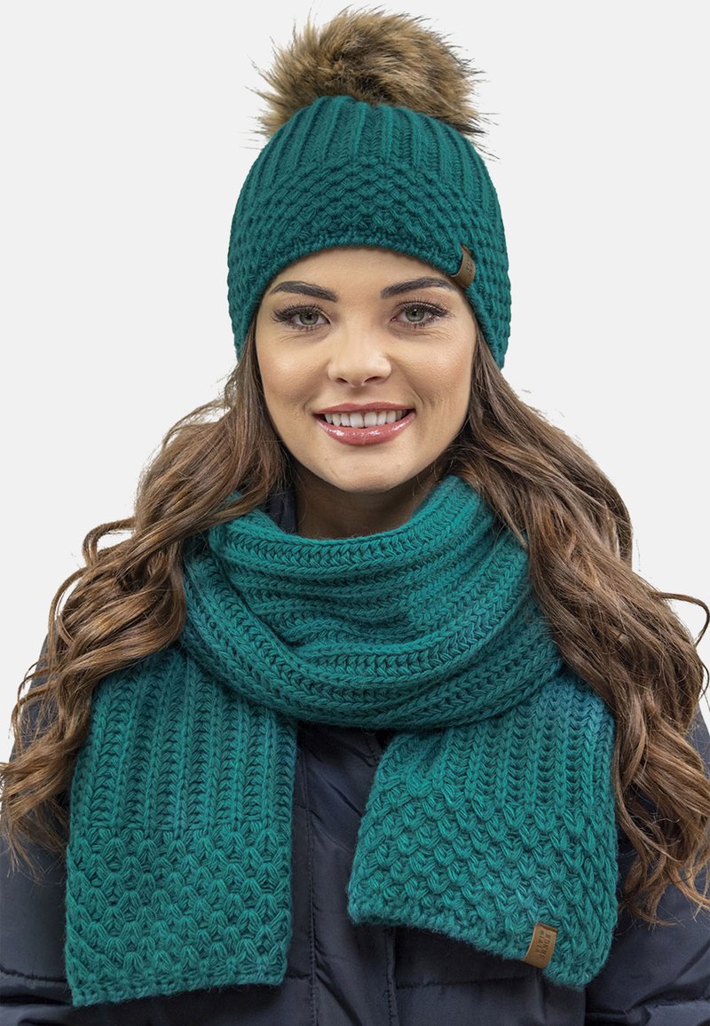Bonnet en tricot teal avec un pompon en fausse fourrure, associé à une écharpe texturée assortie. Les deux présentent des motifs variés et sont fabriqués en laine douce.