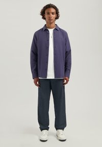 Jonge man met krullend haar, gekleed in een paarse blouse over een wit T-shirt, donkere broek, witte sneakers en een zilveren ketting, die rechtop staat.