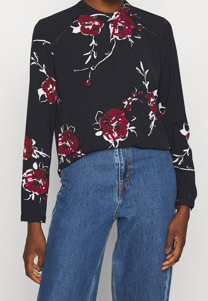 Blouse noire à manches longues avec imprimé floral blanc et rouge, dotée d'un col rond et de détails de couture subtils aux coutures des épaules.