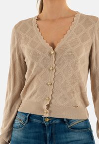 Cárdigan de punto color beige con escote con ondas, patrón de diamantes y seis botones dorados. Dobladillo y puños acanalados, textura ligera.