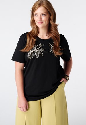 Femme aux longs cheveux roux portant un t-shirt noir à motif floral et un pantalon jaune à jambes larges, debout avec la main dans la poche.