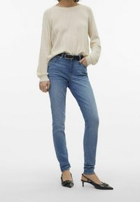 Femme portant un pull en tricot crème, un jean skinny bleu avec une ceinture noire et des talons ouverts noirs avec des nœuds, se tenant devant un fond blanc.