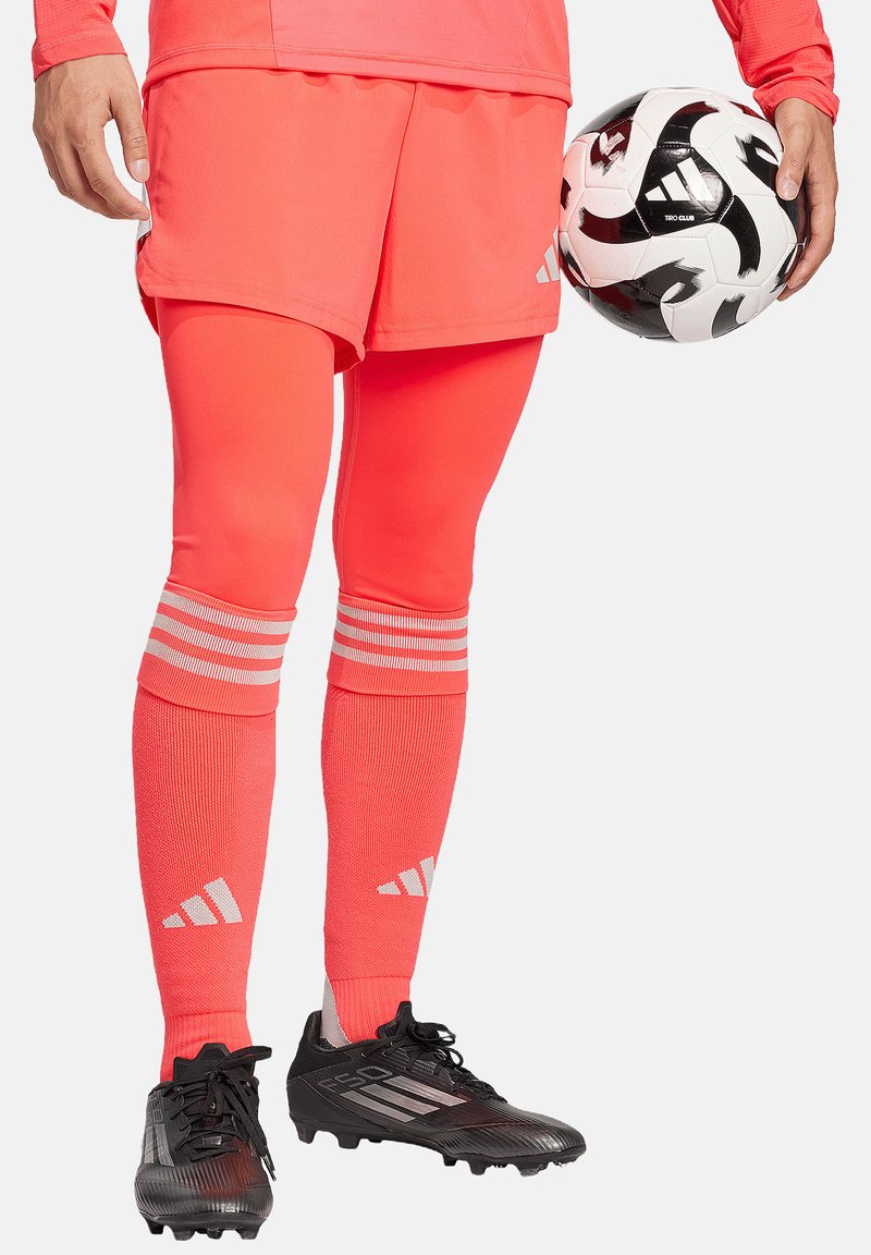 Helder koraalkleurig voetbaluniform met korte shorts en lange sokken met drie witte strepen. Zwarte voetbalschoenen en een zwart-wit voetbals.