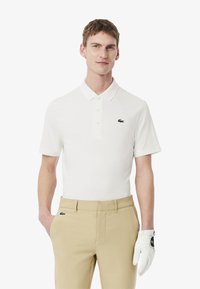 Hvid polo t-shirt med kort ærme, krave og tre knapper, der har et grønt Lacoste-logo. Beigefarvede bukser og en hvid golfhandske er båret.