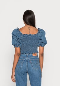 Blouse en jean à manches bouffantes avec un corps smocké et des bords volants ; présente une couleur bleue unie et une coupe ajustée.