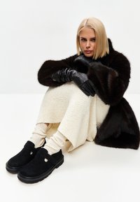Kvinde med blond bob-frisure, der sidder, iført en mørkebrun pelskåbe, sorte læderhandsker, lys strikket nederdel, strømper og sorte slip-on sko.