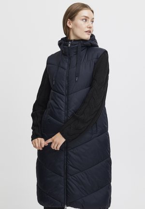 Marineblå puffervest med hette, med quiltet tekstur og svarte cable-knit ermer. Glidelås-lukking og sidelommer gir funksjonalitet.