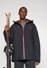 Μαύρο αδιάβροχο μπουφάν σκι με κόκκινο φερμουάρ και λογότυπο, διαθέτει κουκούλα, τσέπες με φερμουάρ και υφή υφάσματος. Σκι κρατάει στο άλλο χέρι.