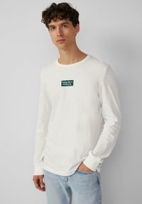 White long-sleeve t-shirt in a light fabric, features a rectangular green label with white text on the chest, paired with light blue jeans.