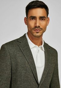 Man met kort, donker haar, gekleed in een donker, gemusterd blazer over een wit polo shirt, die recht in de camera kijkt tegen een effen achtergrond.