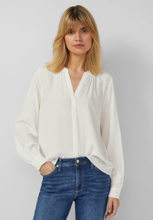 Weiße Bluse mit langen Ärmeln und V-Ausschnitt, geschultert gerafft und aus leichtem Material gefertigt. Getragen mit blauen Jeans und akzentuiert durch ihre entspannte Passform.