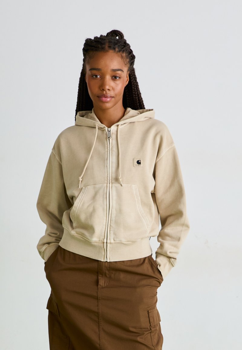 Haut à capuche zippé beige avec un cordon de serrage, une poche kangourou et un petit logo. Porté sur une jupe brune avec des poches latérales. Texture douce.