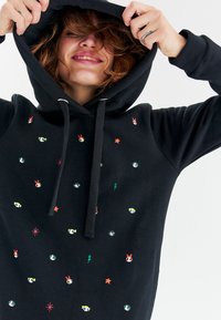 Černá mikina s kapucí a šňůrkou, zdobená barevnými vyšívanými vzory animovaných postav, hvězd a motivy blesků. Měkká textura.