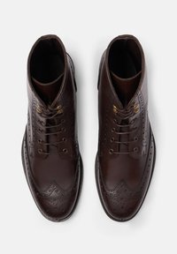 Paio di stivali alla caviglia in pelle marrone scuro lucida con dettagli brogue e lacci, visti dall'alto su sfondo bianco.