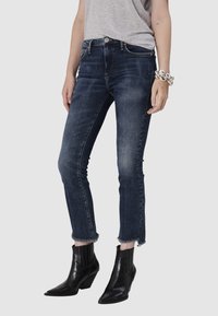 Jean en denim bleu foncé avec un ourlet effiloché, coupe taille mi-haute et détails de décoloration subtils. Associés à des bottines noires à bout pointu.