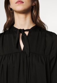 Blusa negra con detalle de lazo en el cuello y diseño plisado. Fabricada con tejido suave, con un corte holgado y ligero brillo.