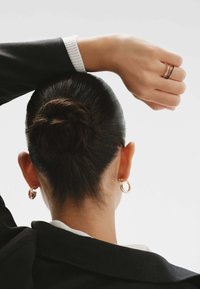 Aros de aro dorados adornan las orejas del modelo. Un elegante moño acentúa el cuello, complementado por una chaqueta negra y un cuello blanco.