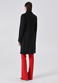 Donna di spalle che indossa un lungo cappotto nero, pantaloni rossi a gamba larga, orecchini a cerchio dorati e scarpe nere con punta su uno sfondo neutro.