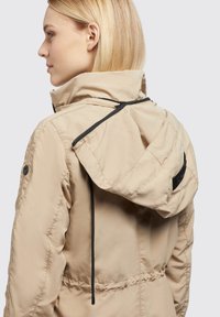 Beige Outdoor-Jacke mit hohem Kragen, verstellbarem Kordelzug und Reißverschlussakzenten. Der Stoff wirkt leicht und glatt.