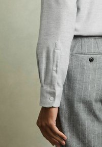 Grå langærmet skjorte med knaplukning ved håndleddet, parret med grå-stribede bukser. Stoffet ser glat og blødt ud.