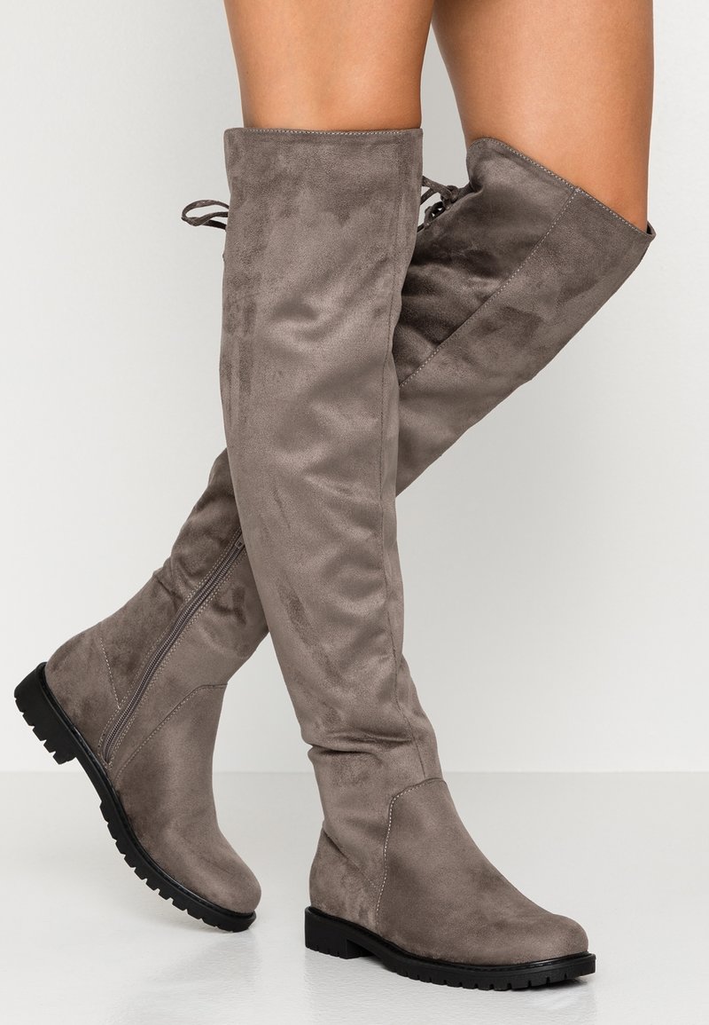 Bottes grises en daim montant au-dessus du genou avec une semelle en caoutchouc plate, dotées d'un bout rond et d'accents de lacets sur le dessus. Détail de fermeture éclair sur le côté.