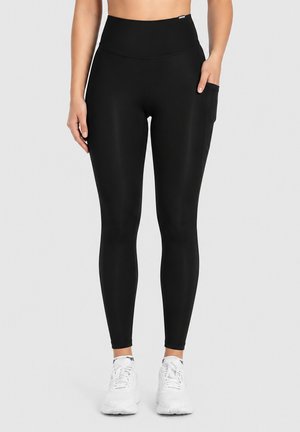 Femme portant des leggings noirs taille haute avec une poche latérale et des chaussures de sport blanches sur un fond uni.